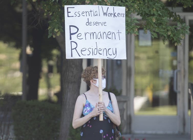 A woman carries a sign that reads: Essential workers deserve permanent residency.